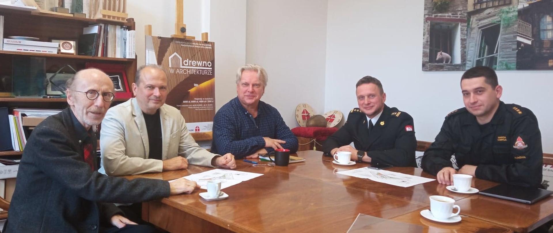 Przy drewnianym stoli siedzi pięciu mężczyzn. Dwóch z nich ubrani sią w mundury strażackie. 