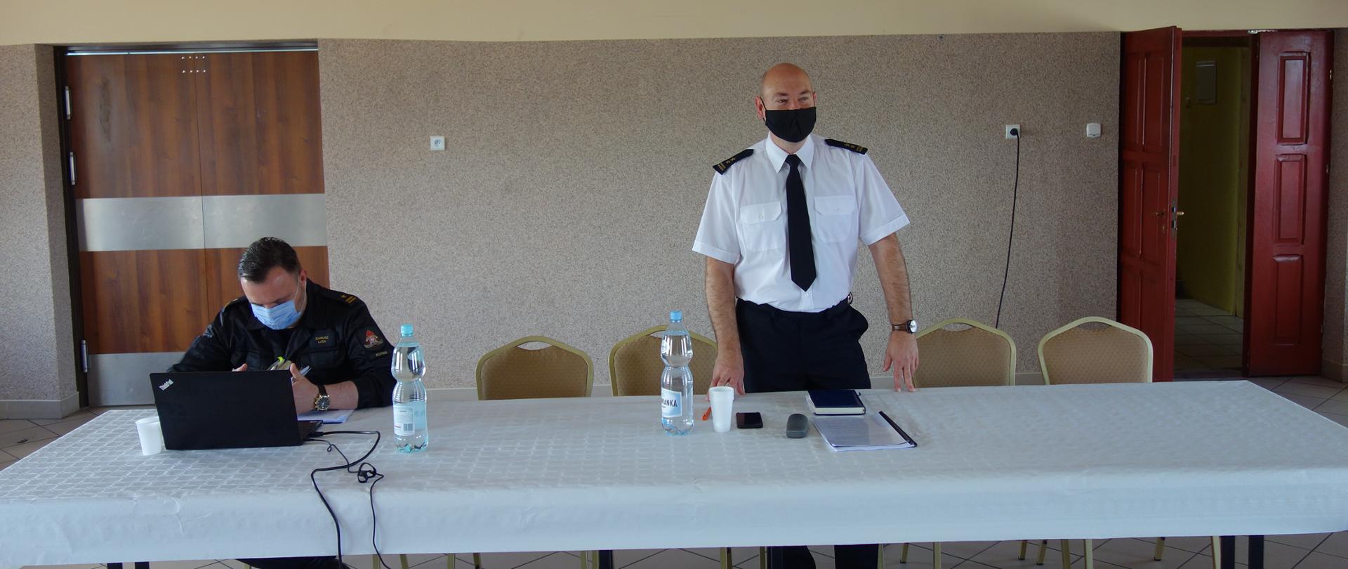 Przy białym stole w sali OSP stojąc przemawia Zastępca Komendanta Państwowej Straży Pożarnej w Kutnie. Z jego prawej strony siedzi Naczelnik wydziału operacyjno szkoleniowego a przed nim laptop. Na stole stoją dwie butelki z wodą i kubki białe.