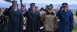 Uczestnicy ceremonii upamiętniającej oddają hołd przy grobie; wśród obecnych znajdują się przedstawiciele służb mundurowych w galowych mundurach, w tym Państwowej Straży Pożarnej, Wojska Polskiego oraz Policji