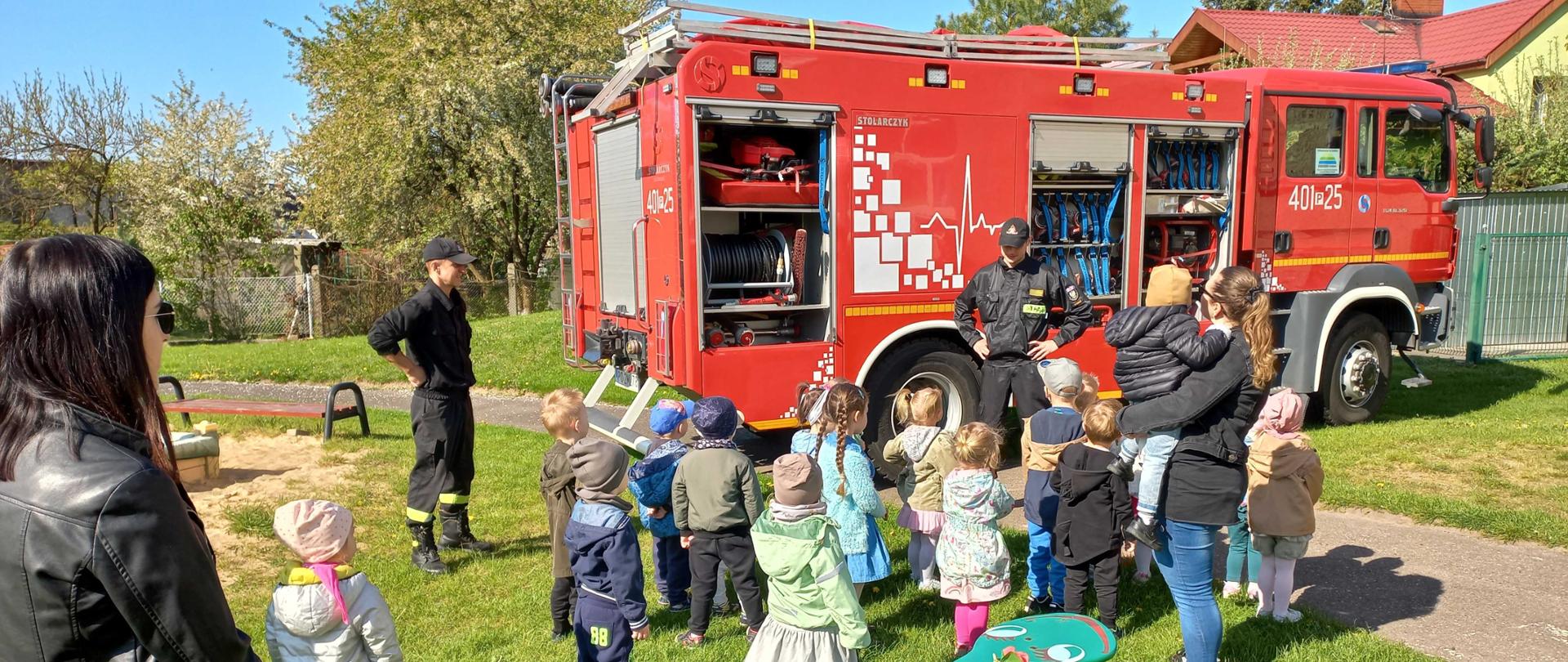 Zdjęcie przedstawia strażaków oraz sprzęt i pojazdy strażackie podczas edukacji dzieci i młodzieży.
W tle budynek drzewa.
