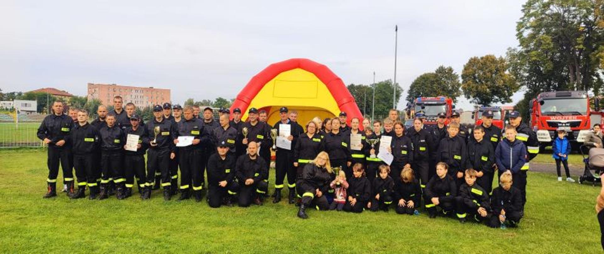 Zdjęcie grupowe. Na środku zdjęcia uczestnicy zawodów. Strażacy i strażaczki ubrani w czarne ubrania koszarowe. Wszyscy uśmiechnięci i zadowoleni z wyników zawodów.