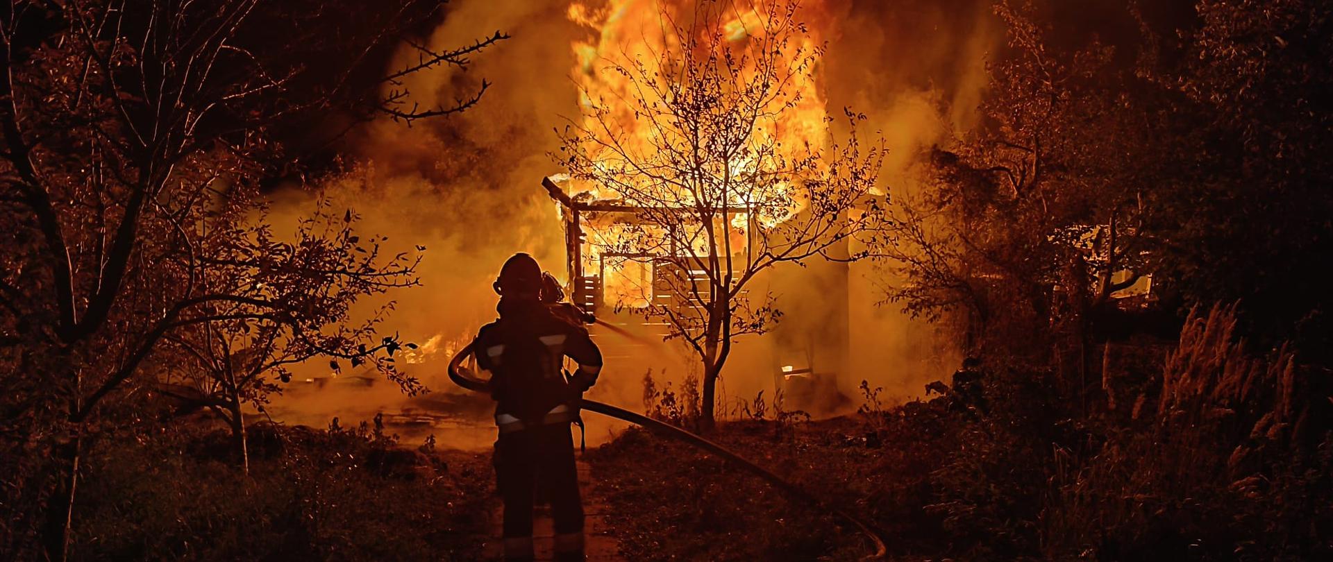 Zdjęcie przedstawia strażaków w trakcie działań gaśniczych. W tle płonąca altana. Na pierwszym planie strażacy podający prąd gaśniczy za pomocą linii wężowej na źródło ognia. Wokół widać krzewy i drzewa z ogródków działkowych.