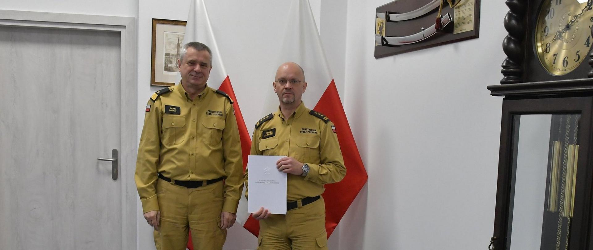Dwóch funkcjonariuszy Państwowej Straży Pożarnej w mundurach koloru musztardowego stoi obok siebie strażak w okularach trzyma białą teczkę za mężczyznami stoją dwie flagi Polski na ścianą wiszą obraz oraz szabla obok ustawiony jest zegar.