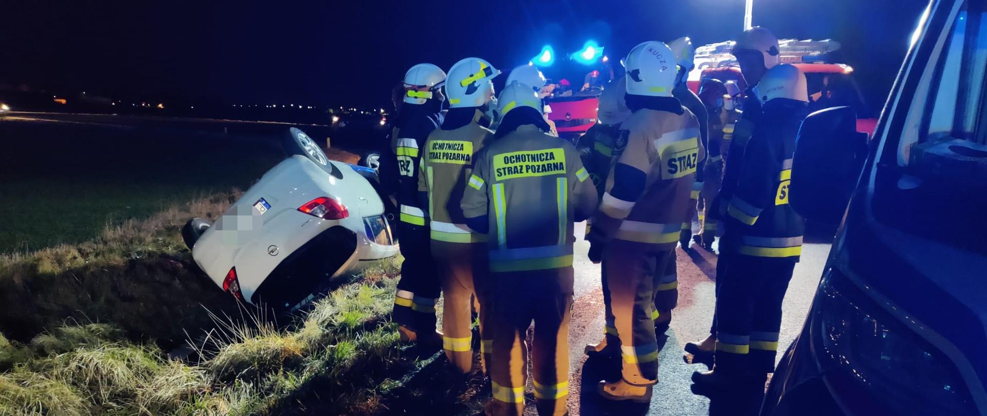 Na zdjęciu samochód osobowy biały w rowie na poboczu stoją strażacy 