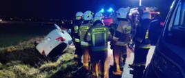 Na zdjęciu samochód osobowy biały w rowie na poboczu stoją strażacy 