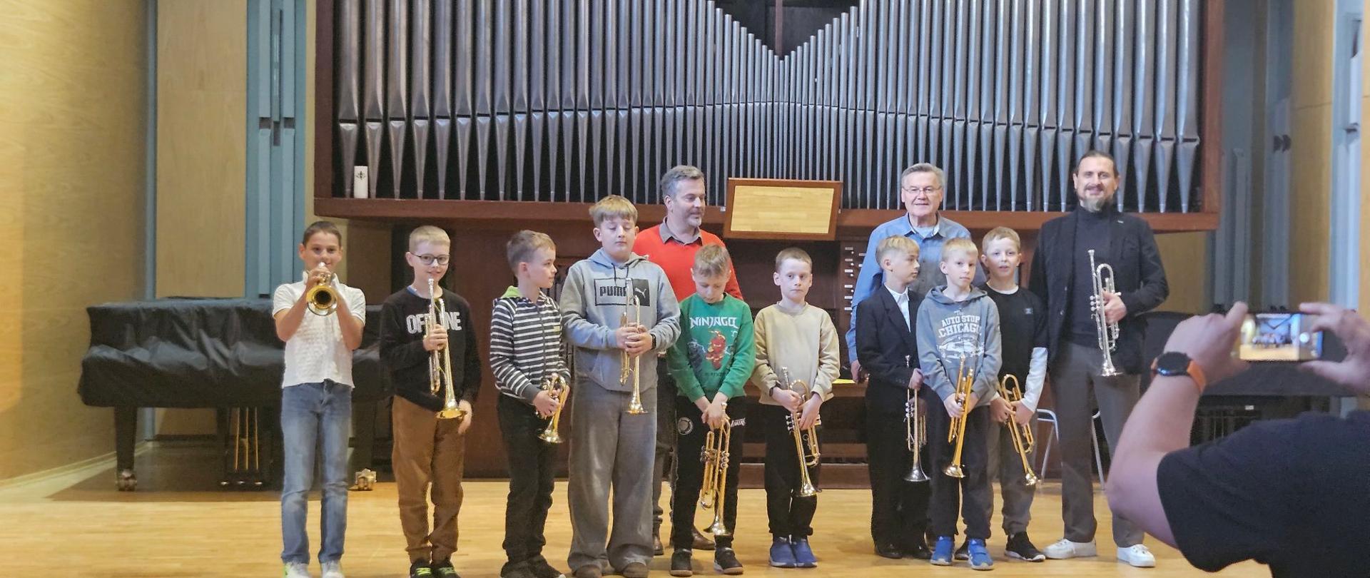 Na zdjęciu uwieczniono grupę młodych muzyków oraz ich instruktorów podczas warsztatów odbywającego się w szkole muzycznej lub sali koncertowej. Pierwszy plan: Dziewięciu chłopców w różnym wieku stoi w rzędzie, trzymając trąbki. Chłopiec po lewej stronie trzyma instrument przy ustach, pozorując grę. Są ubrani w stroje codzienne oraz galowe (jeden z nich ma na sobie garnitur). Drugi plan: Za dziećmi stoi trzech dorosłych mężczyzn. Jeden z nich, po prawej stronie, również trzyma trąbkę. Tło: Wyraźnie widoczne są duże organy piszczałkowe, które dominują w górnej części kadru. Po lewej stronie znajduje się przykryty pokrowcem fortepian. Perspektywa: Zdjęcie jest robione „zza ramienia” innej osoby, której ręce i smartfon widoczne są w prawym dolnym rogu podczas robienia własnej fotografii. Całość ma charakter pamiątkowego zdjęcia po występie lub warsztatach muzycznych.