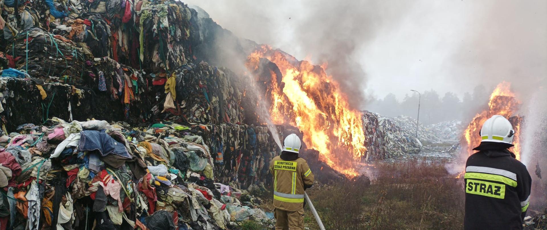 Zdęcie przedstawia dwóch strażaków podających dwa prądy wody na palące się materiały tekstylne. Widoczne są płomienie oraz zadymienie.