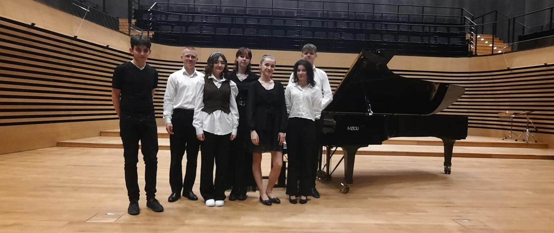 Trzech nastolatków i cztery nastolatki stoją w rzędzie przy fortepianie na estradzie sali koncertowej PSM w Jastrzębiu-Zdroju.