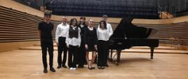 Trzech nastolatków i cztery nastolatki stoją w rzędzie przy fortepianie na estradzie sali koncertowej PSM w Jastrzębiu-Zdroju.
