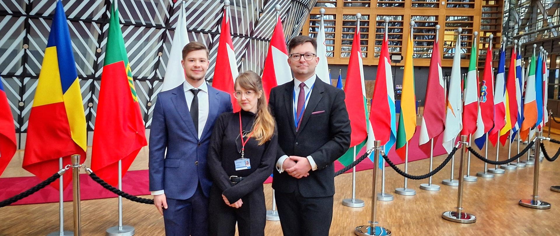 Zdjęcie przedstawia trzy osoby pozujące do zdjęcia w eleganckich strojach biznesowych w reprezentacyjnym holu, w tle znajduje się szereg flag różnych krajów, ustawionych na stojakach i oddzielonych czarnymi barierkami. Mężczyzna po lewej stronie ma na sobie granatowy garnitur, białą koszulę i czarny krawat, kobieta w środku ubrana jest w czarny golf i spodnie, z identyfikatorem na czerwonej smyczy, a mężczyzna po prawej nosi czarny garnitur, białą koszulę, bordowy krawat i okulary. W tle widoczna jest nowoczesna architektura budynku, z drewnianymi elementami i dużą liczbą przeszkleń. Atmosfera sugeruje formalne wydarzenie, prawdopodobnie związane z instytucją międzynarodową lub unijną.