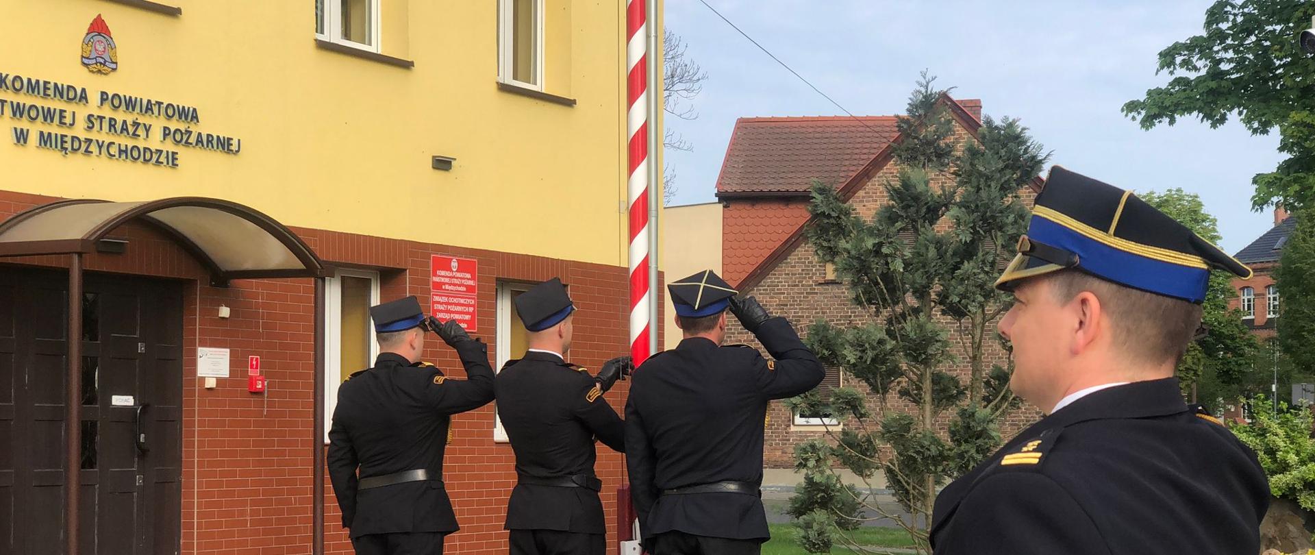 Na pierwszym planie widzimy Zastępcę Komendanta Powiatowego PSP w Międzychodzie, a w tle 3 funkcjonariuszy wchodzących w skład pocztu flagowego podczas podnoszenia flagi państwowej przed masztem. W tle znajduje się budynek KP PSP w Międzychodzie.