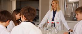 Osoba prowadząca zajęcia stoi przy stole laboratoryjnym, wokół którego znajduje się grupa dzieci w białych fartuchach. Na stole ustawione są kolby, zlewki i probówki z przezroczystą cieczą.