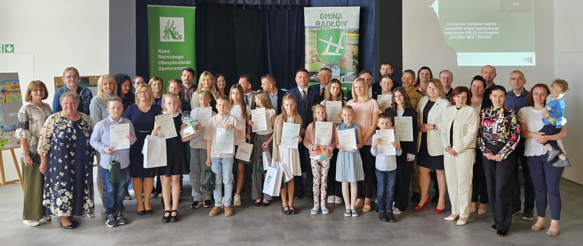 W sali na tle granatowej kurtyny grupa dzieci pozuje do zdjęcia z grupą mężczyzn i kobiet. Wszyscy uśmiechają się do zdjęcia. Z tyłu za nimi zielony plakat z logo KRUS.