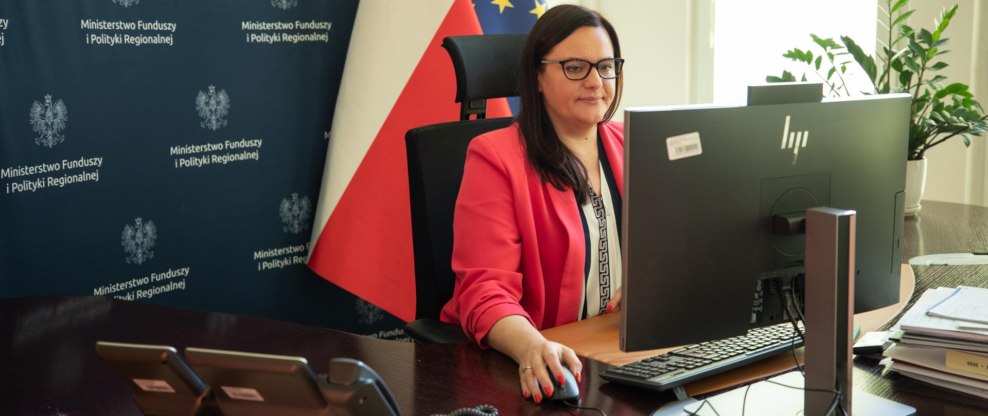 W gabinecie przy biurku z monitorem siedzi wiceminister Małgorzata Jarosińska-Jedynak. Za nią ścianka MFiPR i flagi UE i PL.