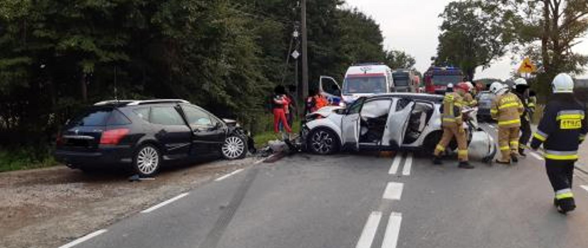 Dwa samochody osobowe po zderzeniu znajdujące się na drodze. Na zdjęciu strażacy prowadzący działania ratowniczo - gaśnicze. W tle widoczna karetka pogotowia, samochód ratowniczo - gaśniczy straży pożarnej oraz dwaj ratownicy medyczni.