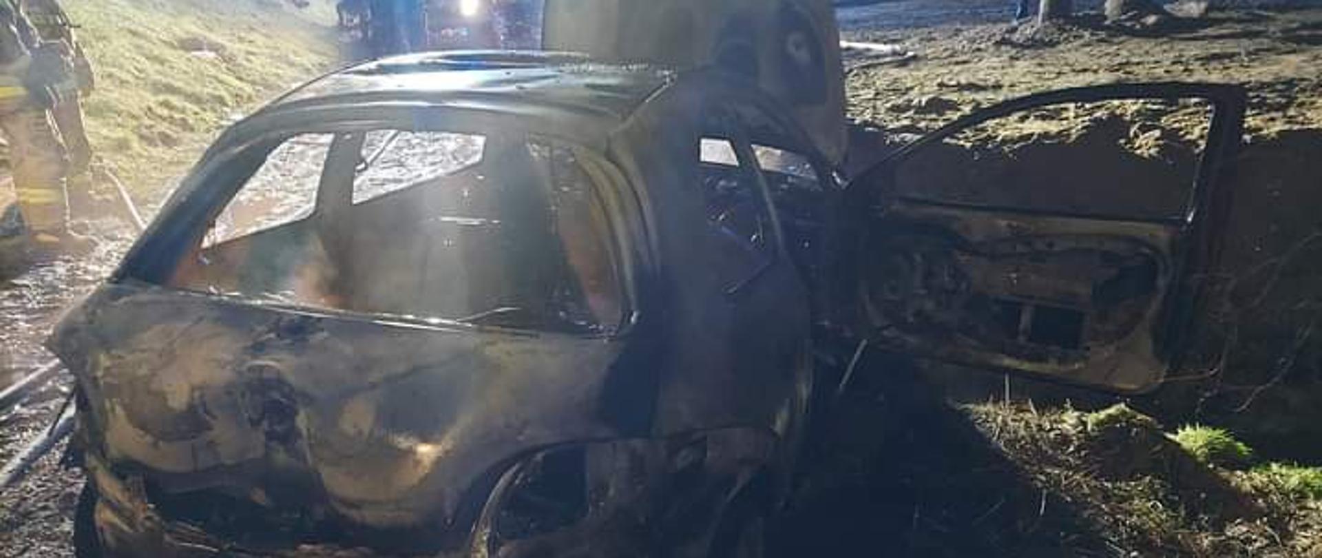 Zdjęcie przedstawia całkowicie spalony samochód osobowy, który zaparkowany jest na drodze gminnej. Samochód ma otwartą maskę oraz drzwi pasażera. Pora nocna. Na zdjęciu widać strażaków oraz samochód strażacki. 
