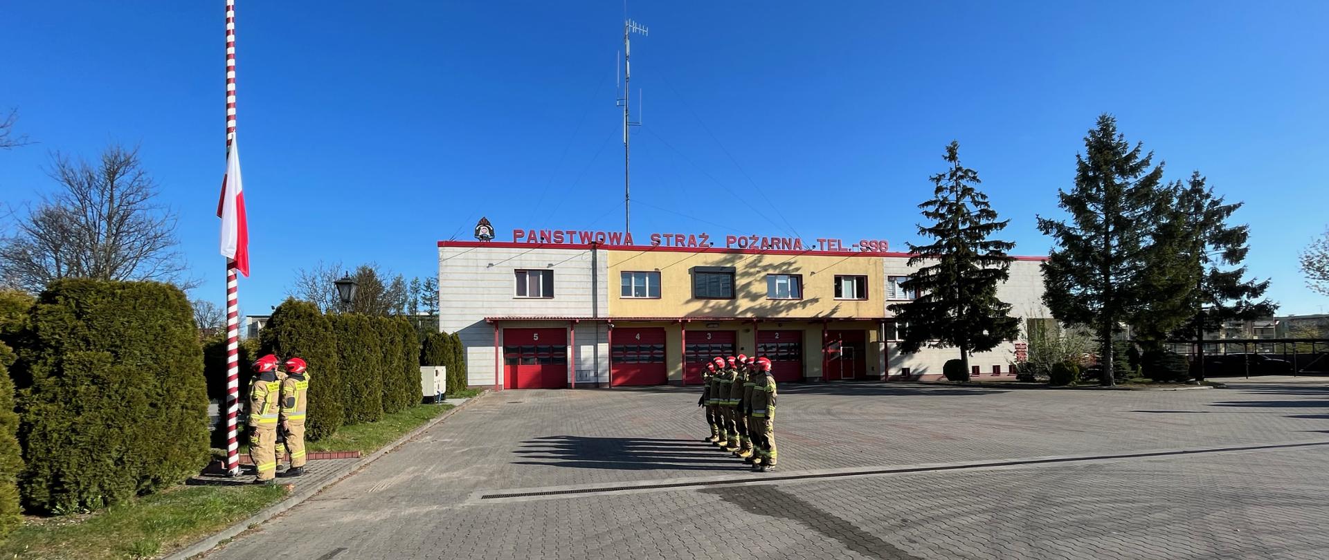 Plac zewnętrzny Komendy Powiatowej PSP w Chojnicach, wyłożony kostką brukową w szarym kolorze. W tle budynek straży pożarnej na tle niebieskiego nieba. W centrum kadru ustawieni w szeregu strażacy oddają honory. Troje strażaków przy maszcie wciąga flagę, która jest widoczna w połowie wysokości masztu. 