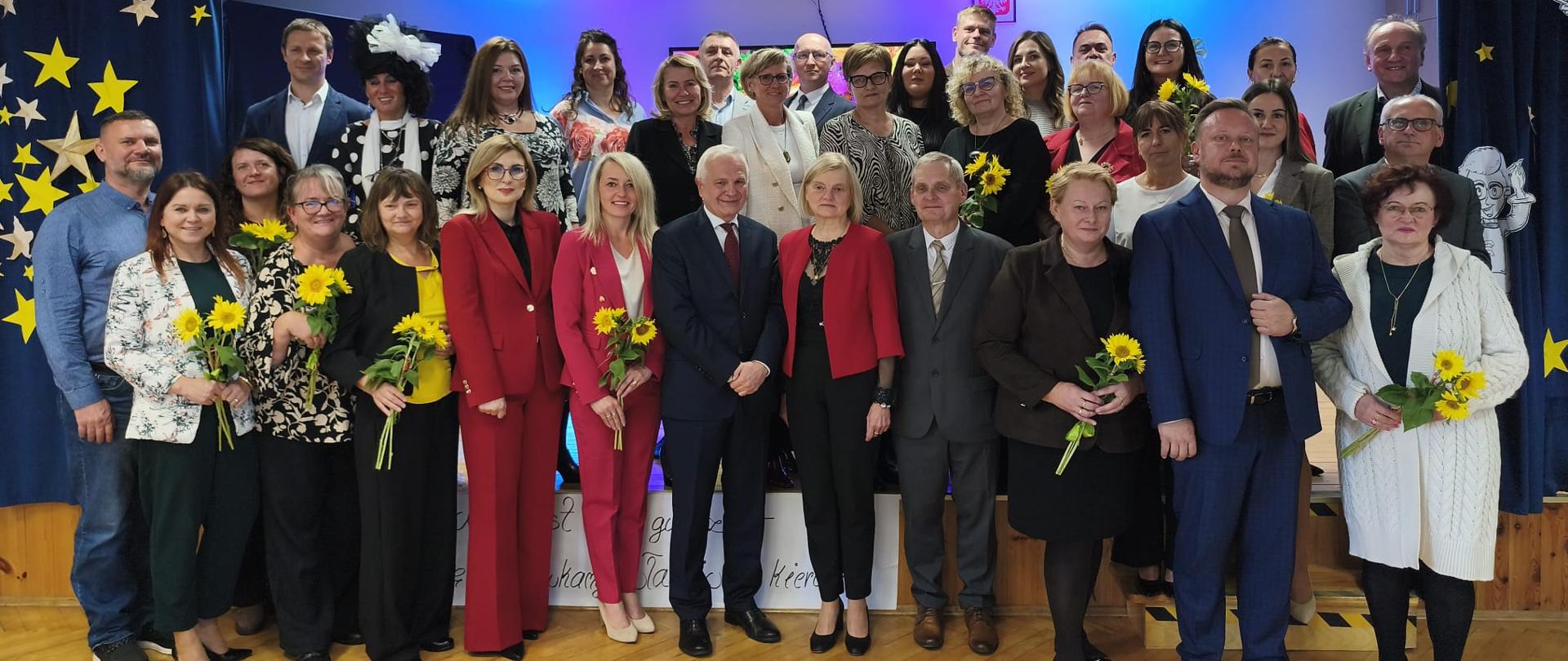 Grupa nauczycieli i pracowników szkoły pozuje do wspólnego zdjęcia przed sceną udekorowaną gwiazdami. Wiele osób trzyma słoneczniki.