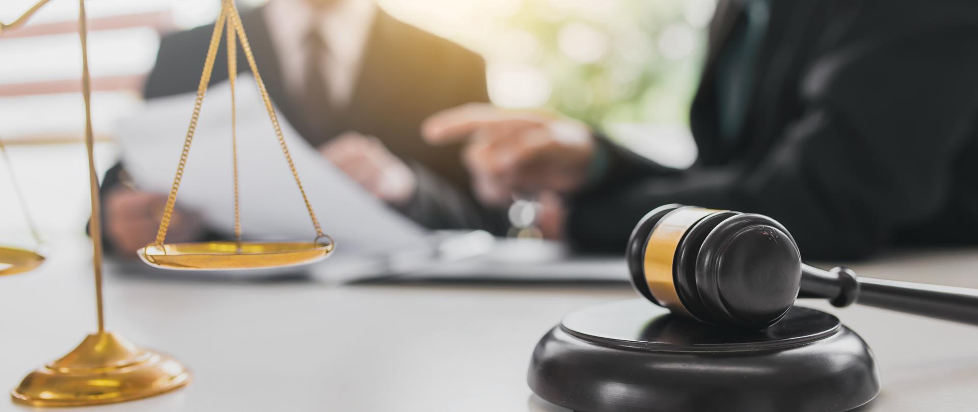 Sędziowski młotek oraz waga szalkowa na tle dwóch dyskutujących ze sobą osób 