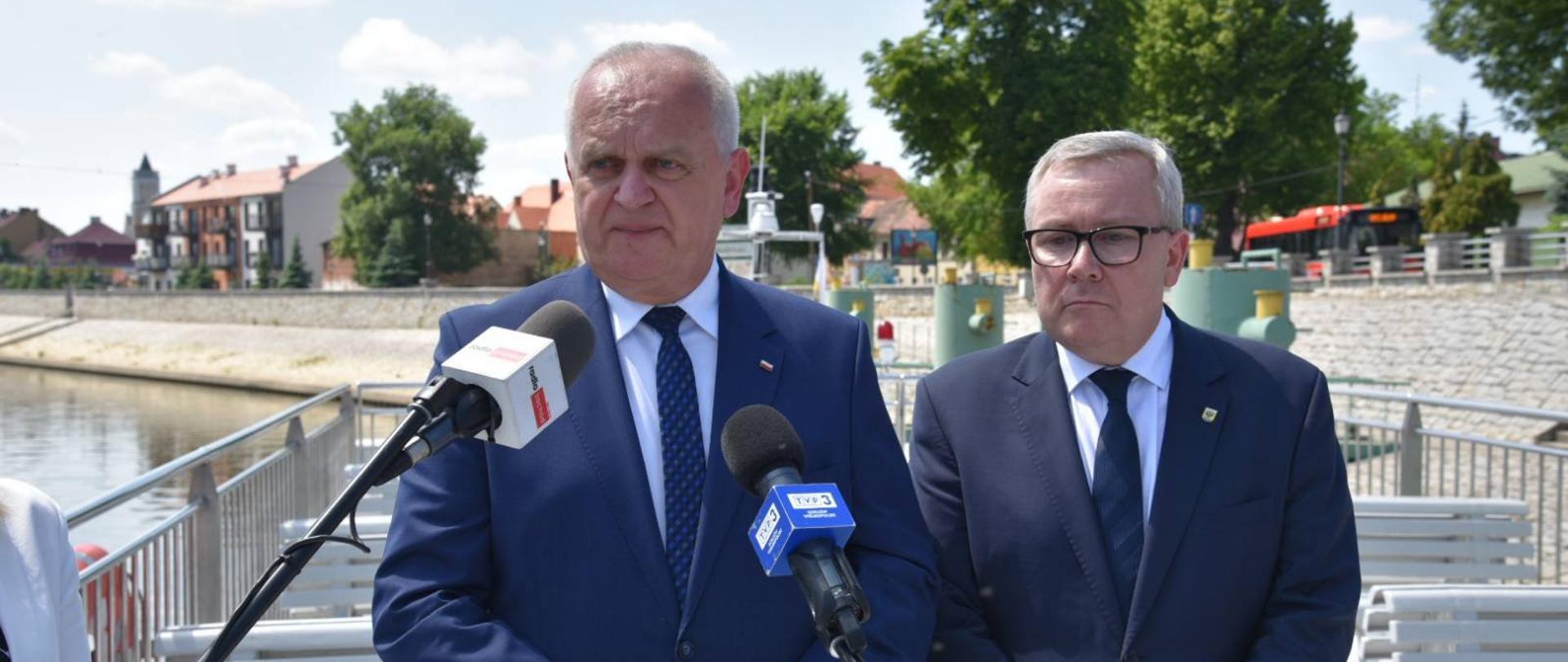 Wojewoda i Prezydent Nowej Soli stoją na przystani, w tle nadbrzeże i rzeka. Przed nimi stoją mikrofony dziennikarzy 