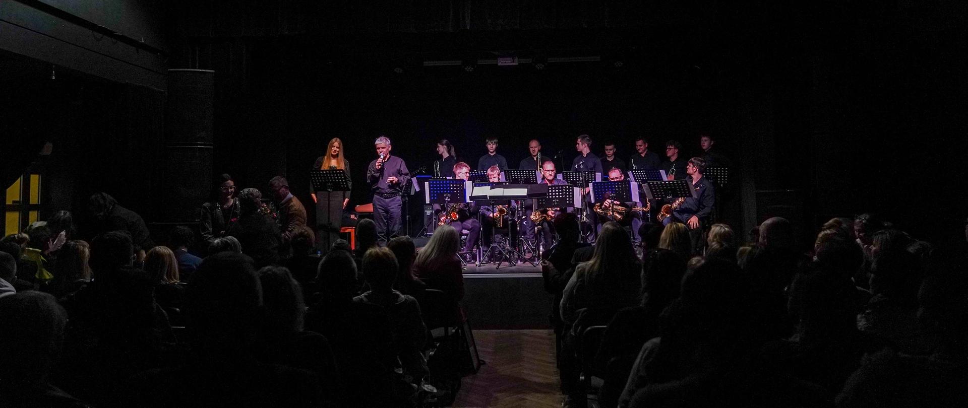 Szerokie ujęcie koncertowe. Na oświetlonej scenie, przed czerwoną kurtyną, stoi Big Band złożony z około piętnastu muzyków grających na instrumentach dętych (głównie saksofonach i trąbkach). Centralnie, tyłem do widza, znajduje się dyrygent, a tuż przed nim, frontem do publiczności, stoi wokalistka w pomarańczowej bluzce, śpiewająca do mikrofonu. Scena oświetlona jest mocnymi reflektorami. Na pierwszym planie widoczne są sylwetki publiczności siedzącej w rzędach, zanurzonej w półmroku.