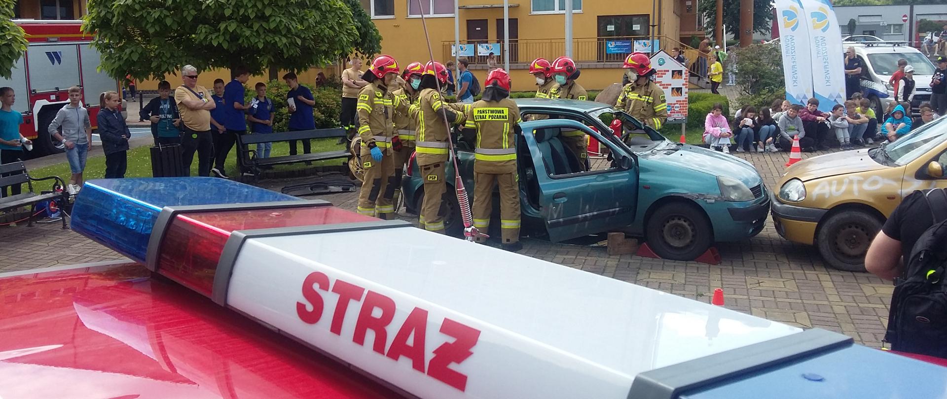Na pierwszym planie belka sygnalizacyjna pojazdu pożarniczego. W tle strażacy w trakcie pokazów ewakuują osobę z uszkodzonego pojazdu.