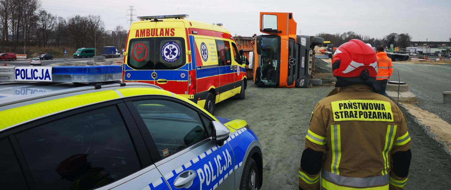Zdjęcie przedstawia strażaka w pisakowym mundurze z napisem "Państwowa Straż Pożarna" stojącym przy radiowozie policyjnym. W tle widać przewróconą ciężarówkę oraz karetkę pogotowia koloru żółtego. 