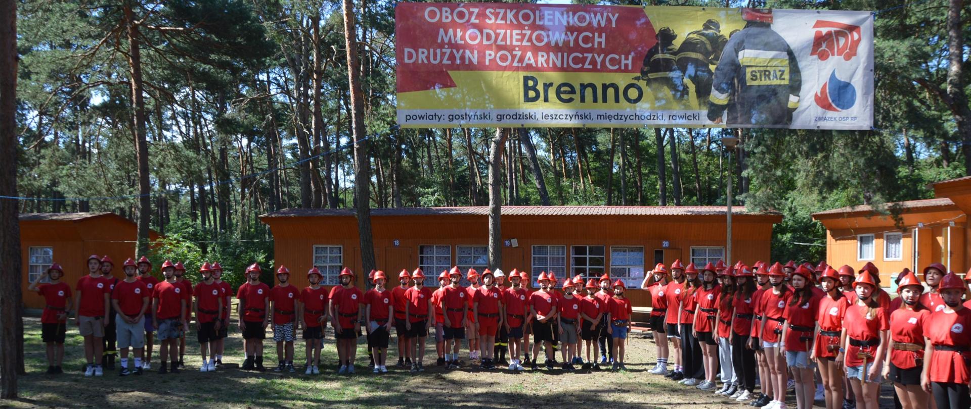 Na placu apelowym odbywa się apel poranny. Poszczególne plutony ustawione są wokół placu w dwuszeregu. Obozowicze mają na sobie czerwone koszulki i hełmy. Nad placem zawieszony jest baner z nazwą obozu, logiem MDP i sylwetkami strażaków. W tle drzewa i domki.