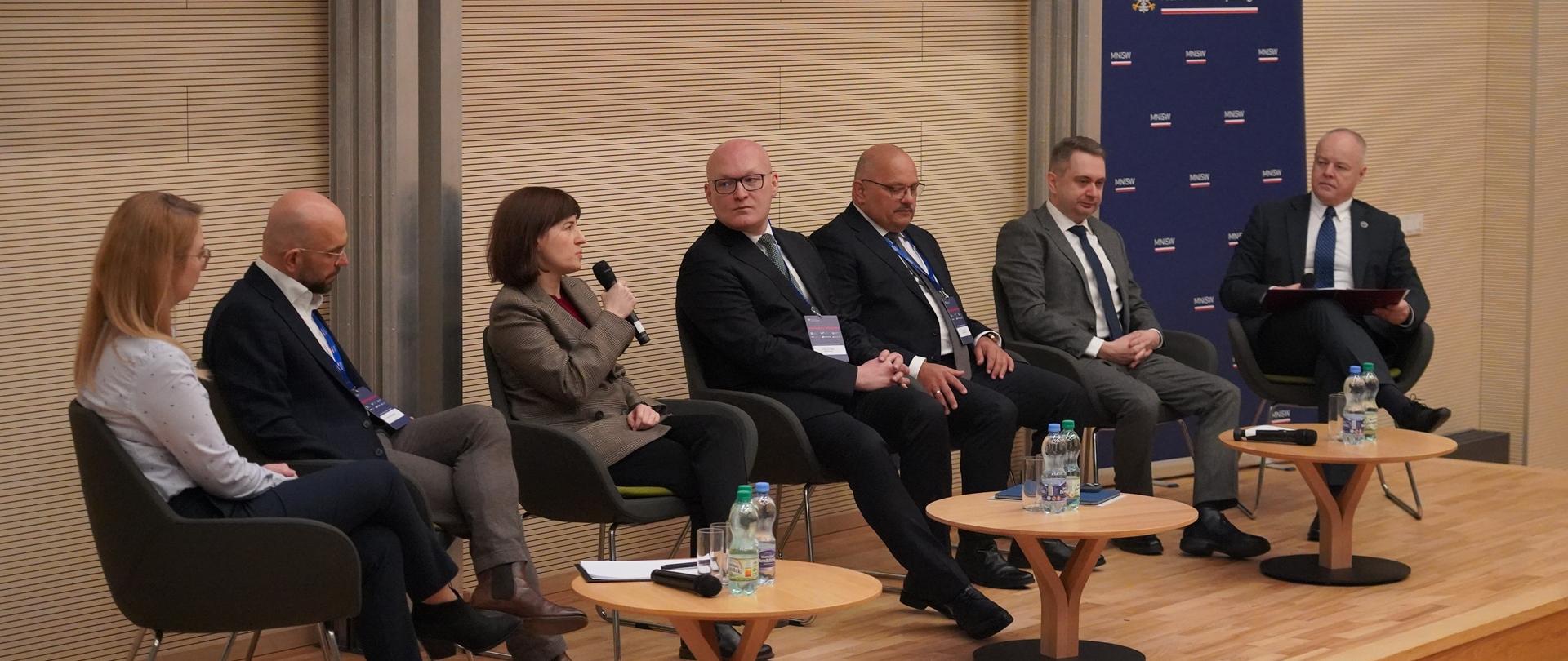 Na podwyższeniu z przodu sali na krzesłach siedzi siedem osób, przed nimi trzy małe okrągłe stoliki, kobieta pośrodku mówi doi trzymanego w ręku mikrofonu, pierwszy z prawej siedzi wiceminister Gzik.