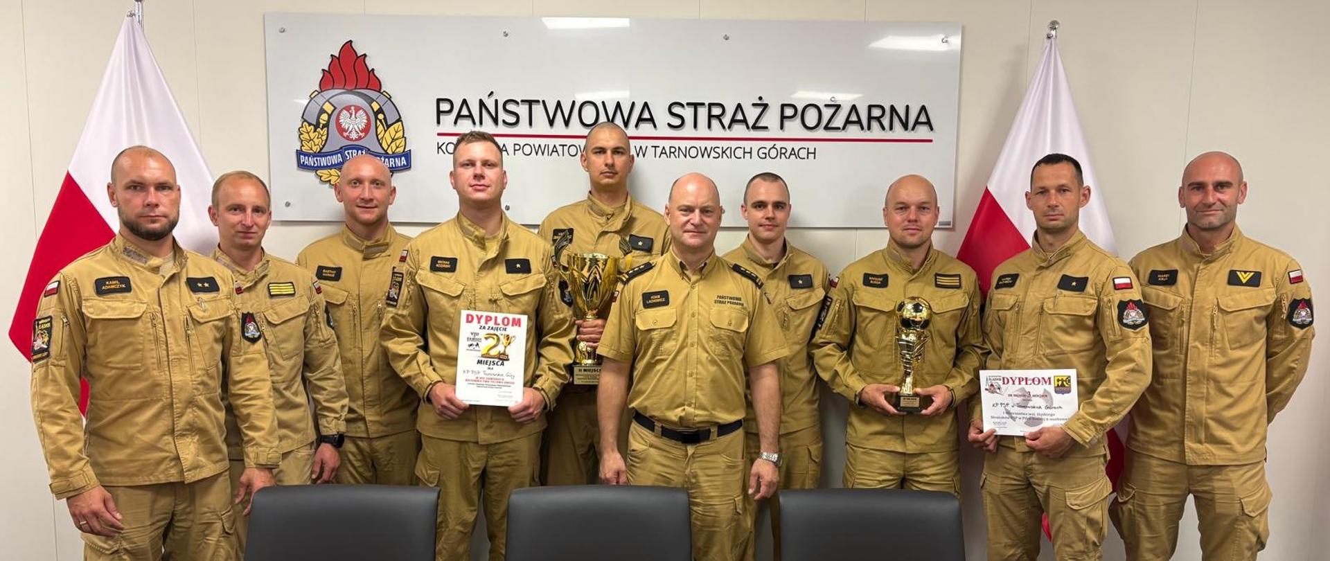 Na zdjęciu widnieje grupa 10 strażaków z Państwowej Straży Pożarnej, pozujących do pamiątkowego zdjęcia w oficjalnym pomieszczeniu.
Oto szczegóły tego, co znajduje się na fotografii:
Tło i otoczenie
Główna tablica: W tle na ścianie wisi duże białe logo oraz napis: „PAŃSTWOWA STRAŻ POŻARNA KOMENDA POWIATOWA W TARNOWSKICH GÓRACH”.
Flagi: Po lewej i prawej stronie kadru ustawione są flagi Polski (biało-czerwone).
Pierwszy plan: Przed strażakami znajduje się długi, drewniany stół konferencyjny oraz czarne, skórzane fotele biurowe.
Strażacy i zdobyte nagrody
Mężczyźni są ubrani w piaskowe (khaki) mundury służbowe i operacyjne PSP. Wygląda na to, że świętują sukces sportowy lub zawodowy, ponieważ trzymają w rękach trofea:
Dyplomy: Dwóch strażaków trzyma w rękach oficjalne dyplomy (na jednym z nich widoczny jest napis „DYPLOM ZA ZAJĘCIE 2 MIEJSCA”).
Puchary: W rękach mężczyzn widoczne są również dwa puchary – jeden klasyczny, wysoki i złoty, a drugi z elementem przypominającym złotą piłkę.
Dowódca/Oficer: Mężczyzna stojący niemal w samym środku wyróżnia się koszulą z krótkim rękawem (również w kolorze piaskowym) oraz dystynkcjami oficerskimi na pagonach.