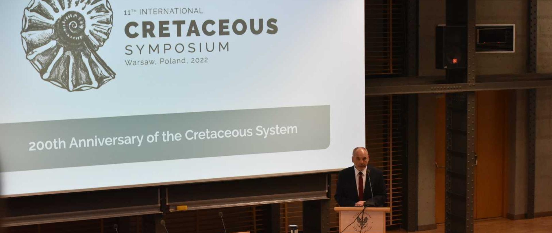 Wiceminister Piotr Dziadzio przy mównicy, za nim napis w języku angielskim: 11 Międzynarodowe Sympozjum Kredowe w Warszawie