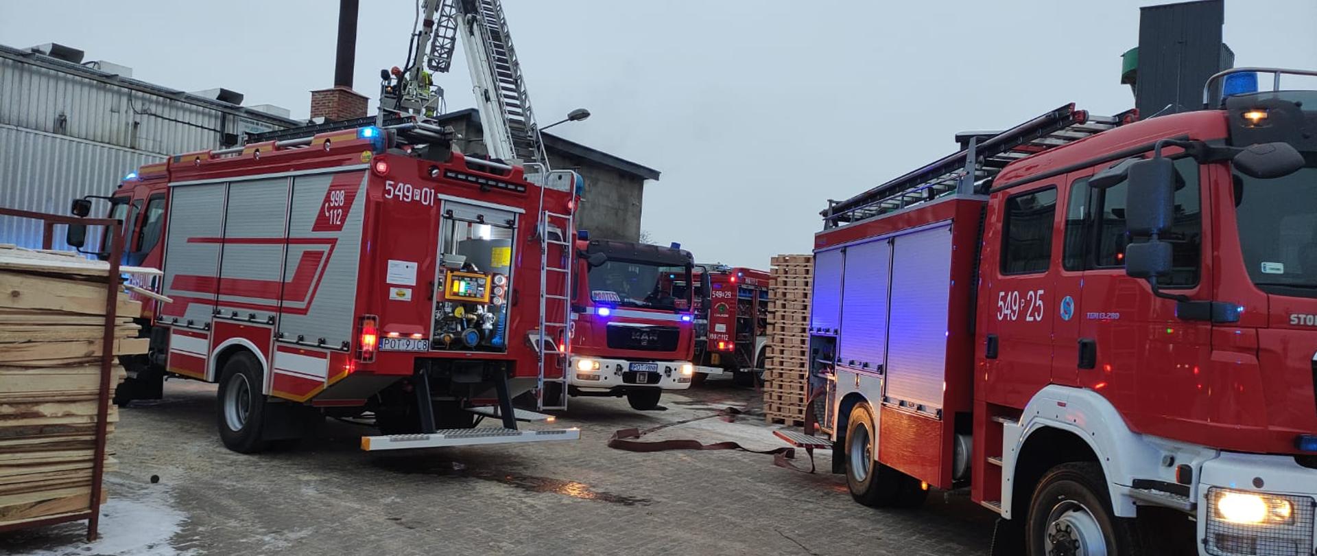 Wozy strażackie stoją pomiędzy drewnianymi paletami i półproduktami drewnianymi na placu. W dalszym planie widać palący się obiekt.