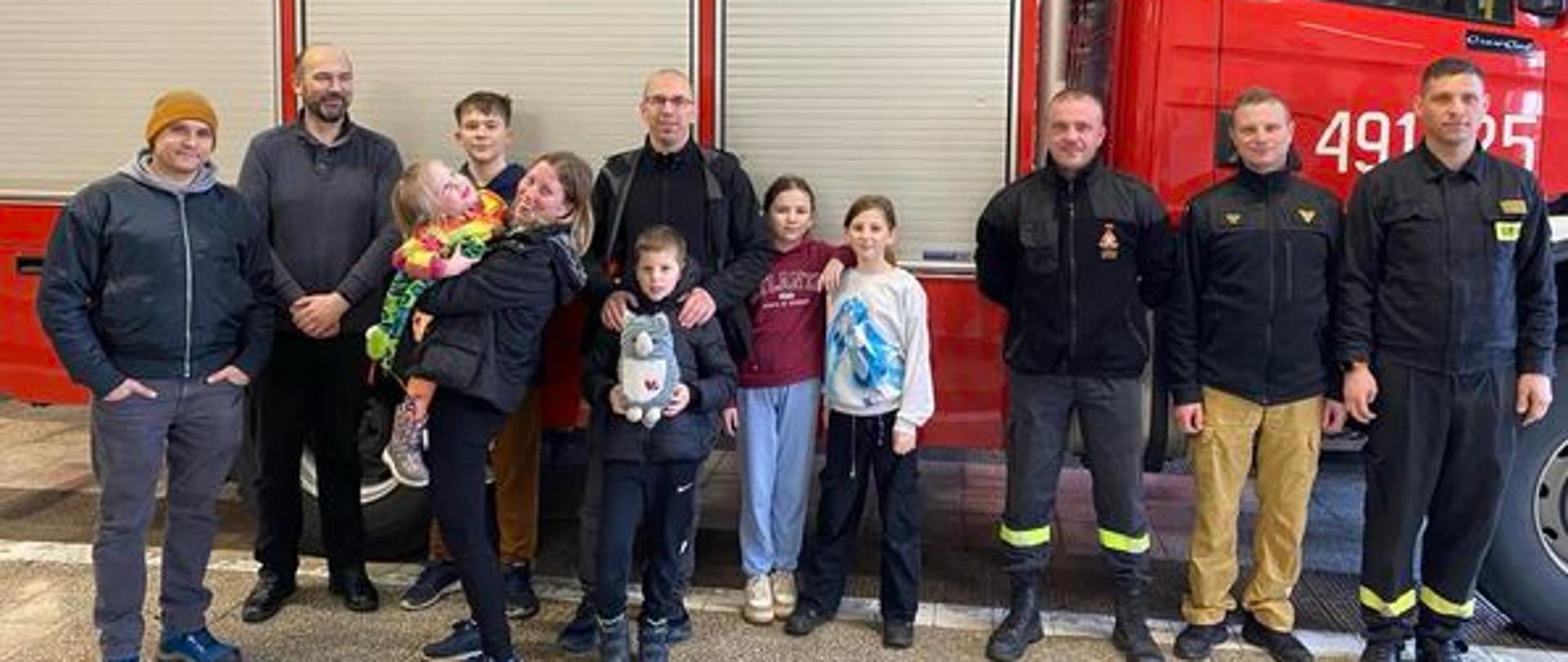 Na zdjęciu znajdują się strażacy ubrani w umundurowanie służbowe oraz osoby cywilne. W tle widać samochód pożarniczy