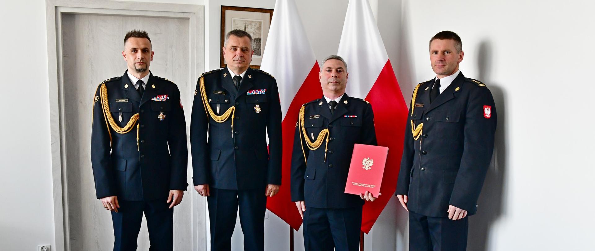 Czterech funkcjonariusze Państwowej Straży Pożarnej stoi przed flagami. Jeden z nich trzyma czerwoną teczkę. 