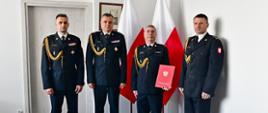 Czterech funkcjonariusze Państwowej Straży Pożarnej stoi przed flagami. Jeden z nich trzyma czerwoną teczkę. 