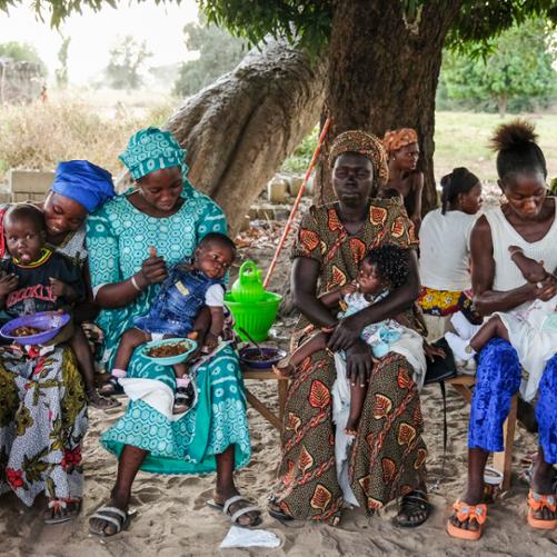 Pod drzewem na ławkach siedzą kobiety i karmią albo przytulają trzymane na kolanach dzieci