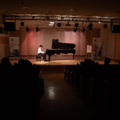 Zdjęcie przedstawia innego ucznia młodszej klasy grającego na fortepianie na scenie sali koncertowej, ujęcie z perspektywy tyłu widowni, widoczne banery szkoły, plakat koncertu oraz dekoracja kwiatowa