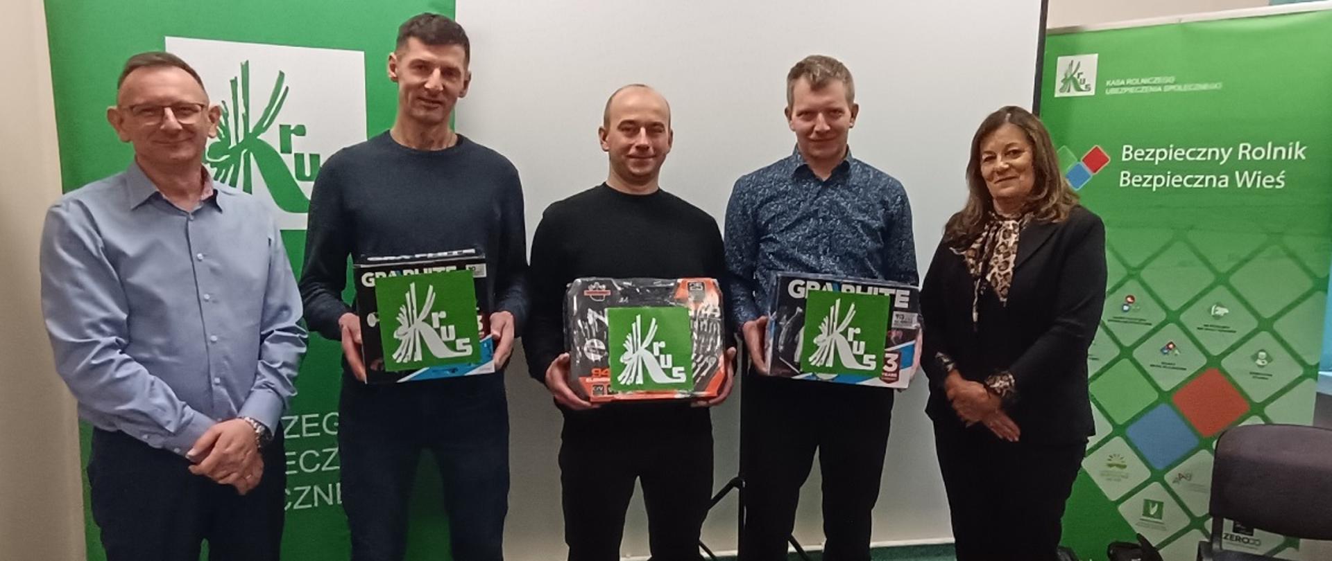 Czterech mężczyzn i jedna kobieta pozują do zdjęcia, uśmiechają się, trzech mężczyzn stojących w środku trzyma pudełka z nagrodami z widocznym na nich logo KRUS. Za nimi znajduje się biały ekran, a po obu jego stronach znajdują się zielone roll-upy z logo KRUS, na tym z prawej strony widoczny jest napis: Bezpieczny Rolnik Bezpieczna Wieś.