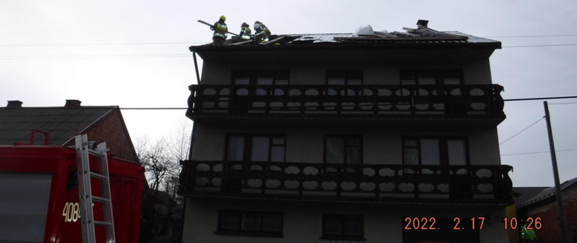 Zdjęcie przedstawia murowany dwupiętrowy budynek na którym doszło do częściowego zerwania dachu. Po lewej stronie widać cześć pojazdu pożarniczego. Na dachu budynku widać czterech strażaków którzy zabezpieczają zerwany dach. Po ich prawej stronie widać część uszkodzonego pokrycia dachowego.