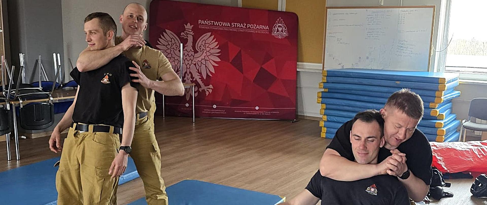 „Strażacy PSP podczas ćwiczeń z samoobrony na sali treningowej wykonują techniki obezwładniania – dwóch uczestników prezentuje chwyt na szyję w pozycji stojącej, a druga para ćwiczy podobną technikę w pozycji siedzącej na matach, pod okiem instruktora.”