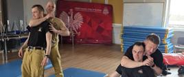 „Strażacy PSP podczas ćwiczeń z samoobrony na sali treningowej wykonują techniki obezwładniania – dwóch uczestników prezentuje chwyt na szyję w pozycji stojącej, a druga para ćwiczy podobną technikę w pozycji siedzącej na matach, pod okiem instruktora.”