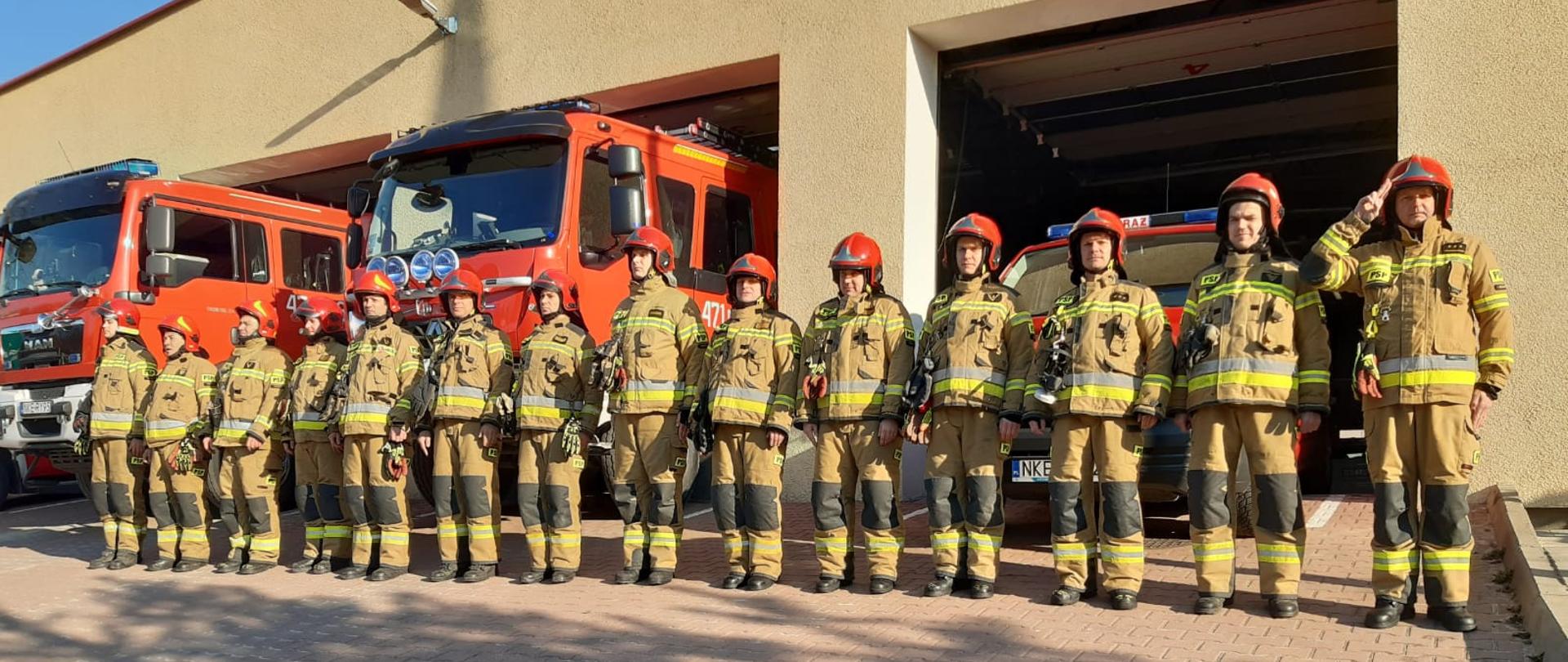 Strażacy ubrani w piaskowe ubrania bojowe ustawieni w szeregu oddają honor poległym strażakom z Ukrainy. Ustawieni w szeregu przed budynkiem komendy, za strażakami stoją czerwone samochody.