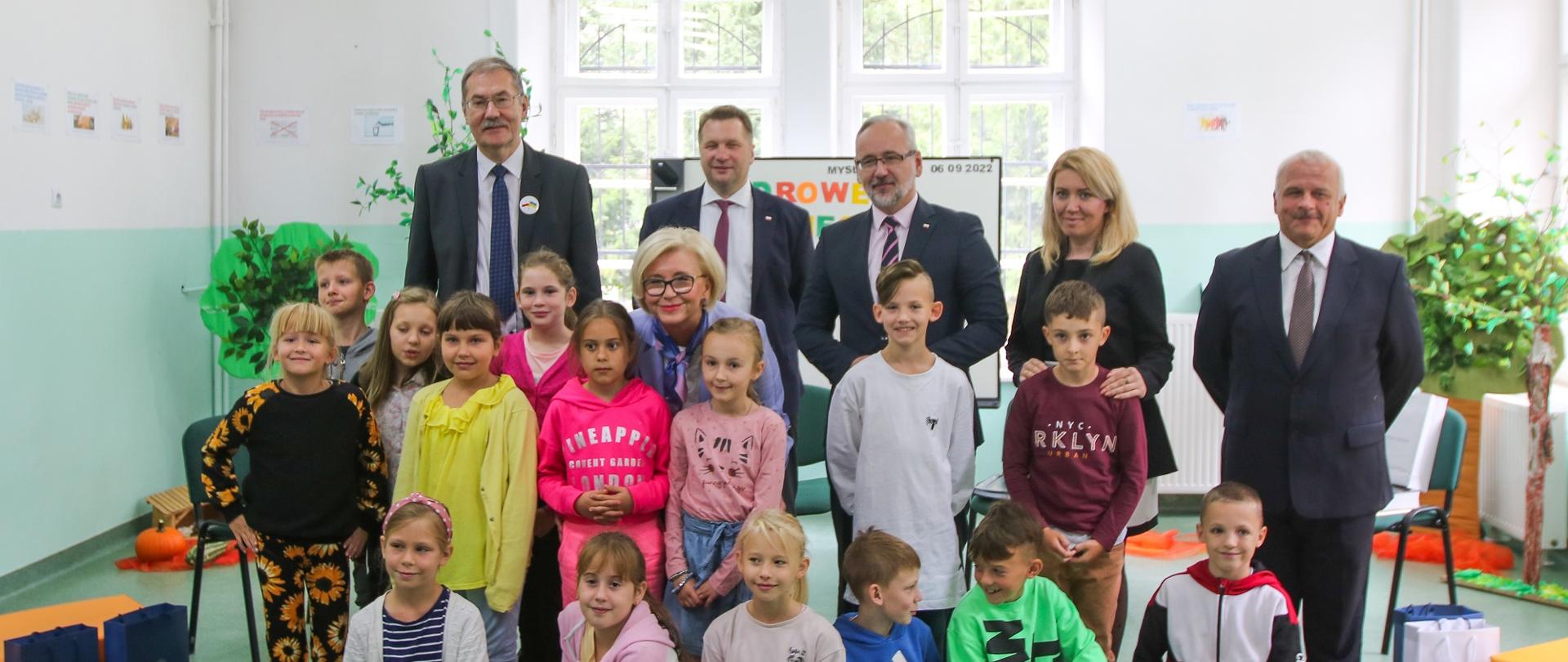 Zdjęcie zbiorowe, grupa dzieci siedzi a za nimi stoją uczestnicy w garniturach, w tle okno.
