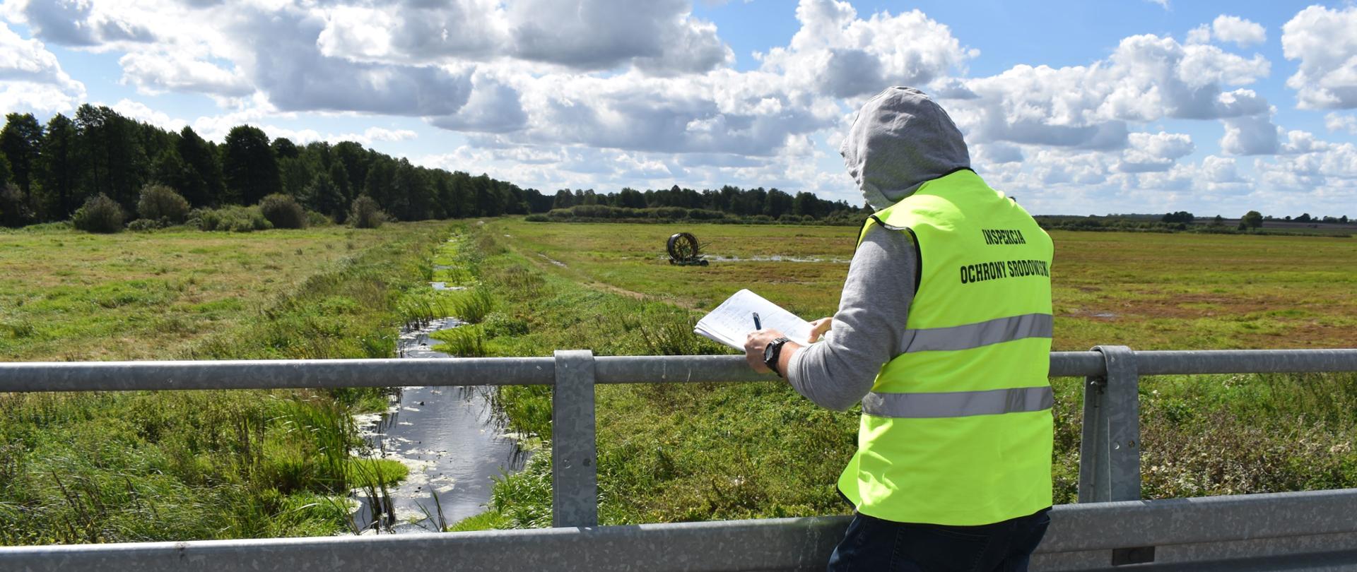 Inspektor Wojewódzkiego Inspektoratu Ochrony Środowiska w Warszawie opierając się o szarą, metalową barierkę mostu prowadzi czynności kontrolne. Kilka metrów pod mostem płynie szeroka na ok. 1 metr rzeka. Po jej obu stronach znajdują się zielone łąki.