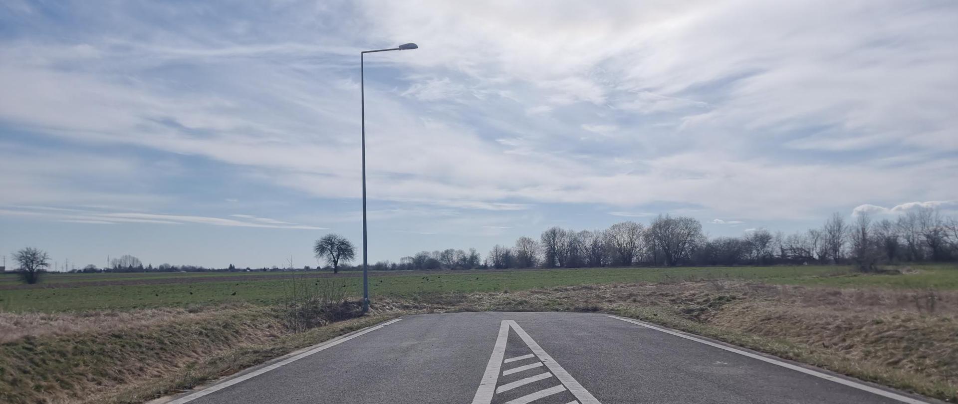 Odcinek drogi z pasem wyłączonym z ruchu na tle pól i nieba.