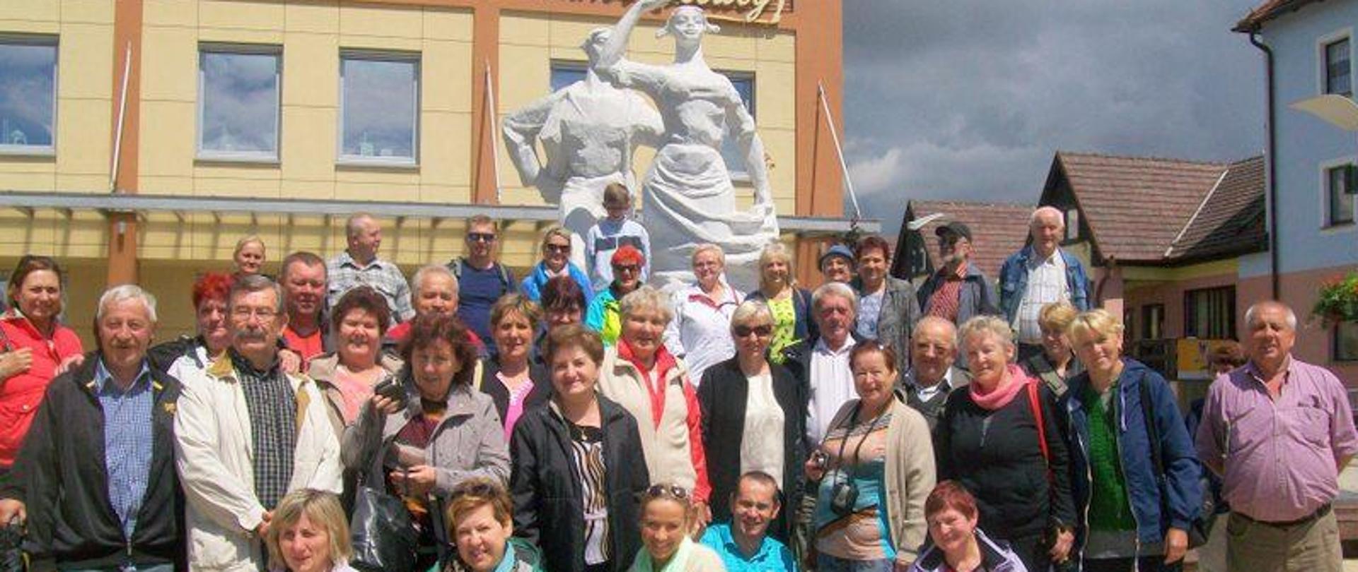 Na zdjęciu widzimy kilkadziesiąt osób mężczyzn i kobiet pozujących do zdjęcia. Pierwszy rząd przysiadł aby nie zasłaniać pozostałych. Grupa stoi przed białym pomnikiem. Dalej w tle widzimy budynek z żółtą o pomarańczową elewacją, do góry napis w kolorze żółtym Gminne Centrum Kultury. Po prawej stronie kadru widzimy budynki mieszkalne, niebo pochmurzone. Grupa stoi na placu utwardzonym żółtą kostką brukową.