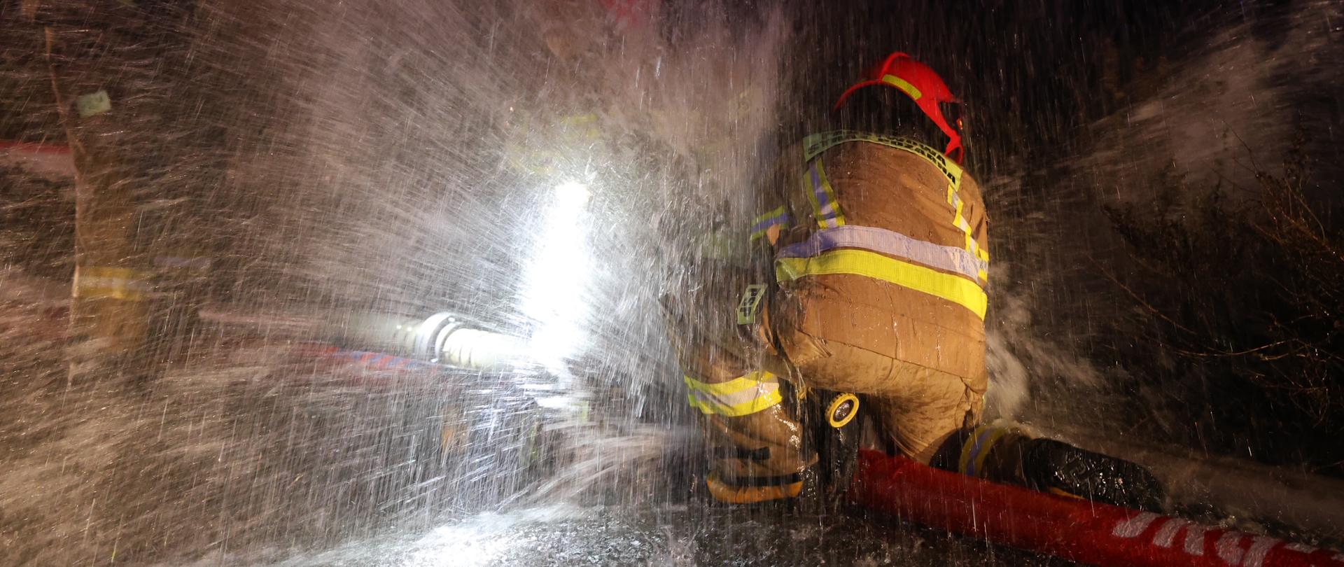 Trzech strażaków próbuje złączyć węże podczas strumienia wody. Jest noc.