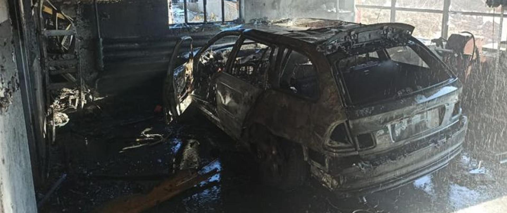 Zdjęcie wykonane po akcji gaśniczej. Centralny punkt stanowi spalony samochód kombi. Auto ugaszone, spalone całkowicie. Auto stoi wewnątrz garażu. Garaż okopcony, ma spalone wnętrze.