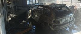 Zdjęcie wykonane po akcji gaśniczej. Centralny punkt stanowi spalony samochód kombi. Auto ugaszone, spalone całkowicie. Auto stoi wewnątrz garażu. Garaż okopcony, ma spalone wnętrze.