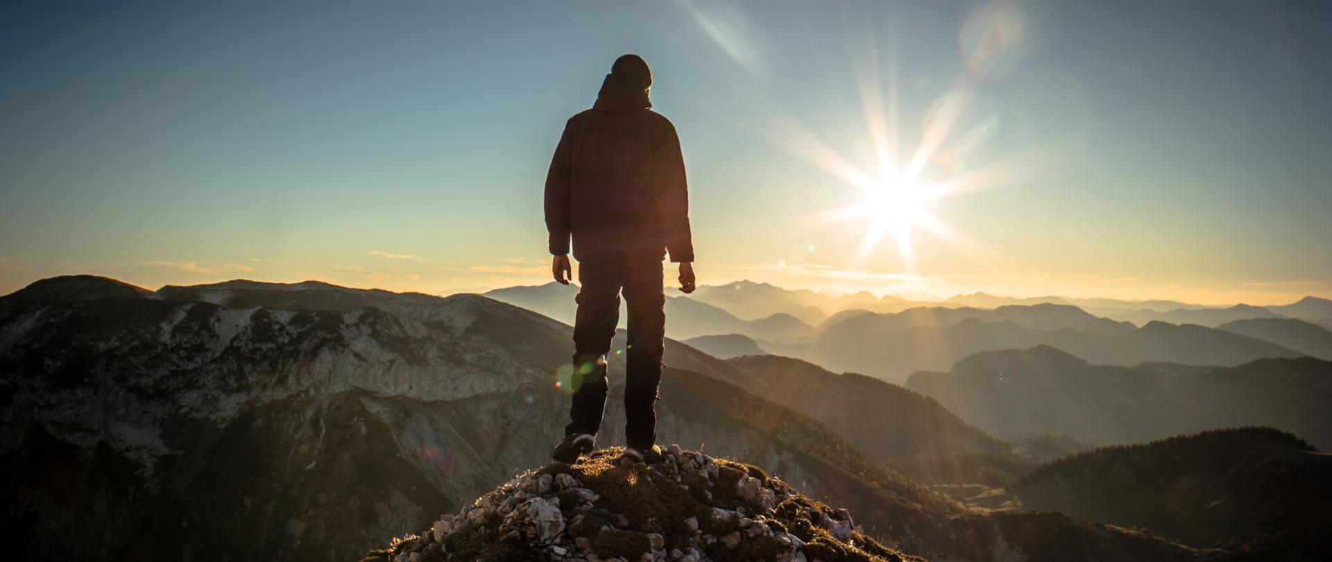 Mężczyzna stojący na szczycie góry, w tle zachodzące słońce.